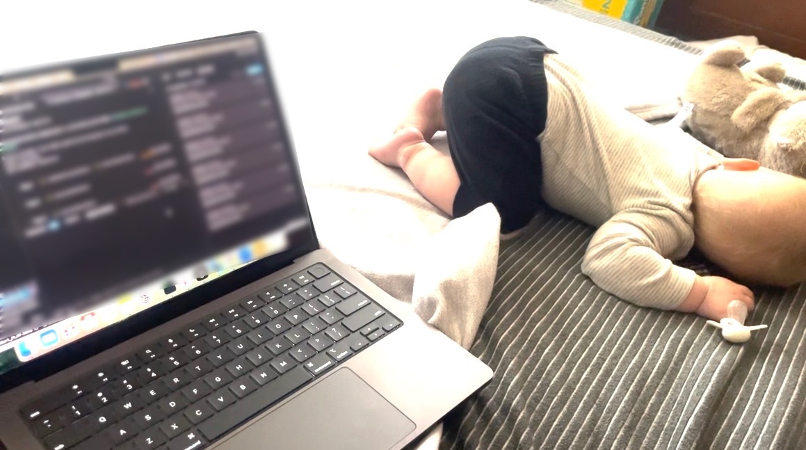 A laptop open on a bed, next to a baby asleep on their front with a pacifier and a stuffed animal.